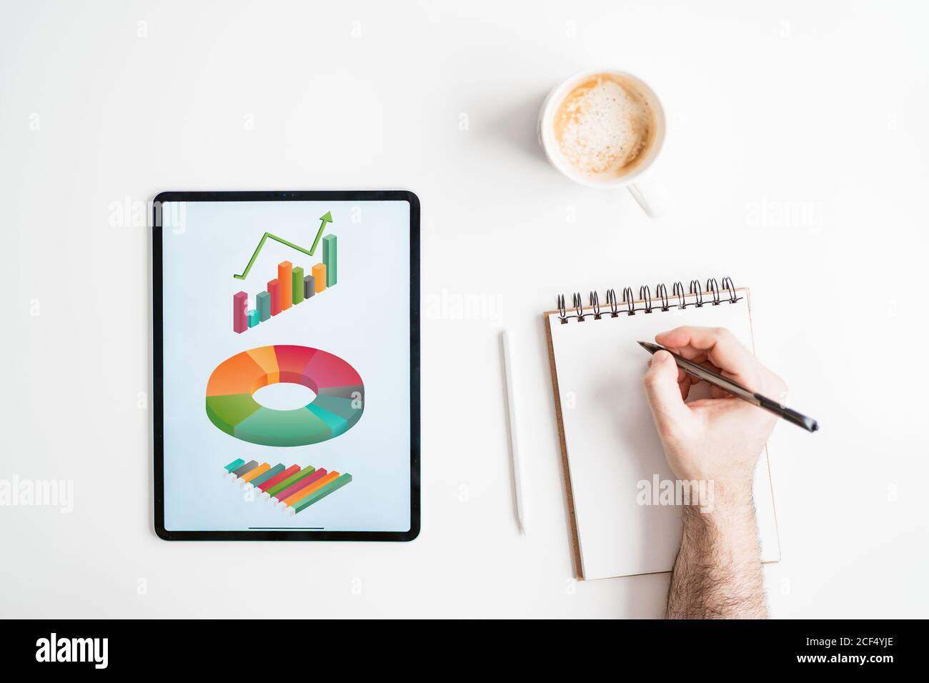 Ein anonymer Mann schreibt in einem Notizbuch. Der Mann arbeitet von zu Hause aus. Das Tablet-Telefon zeigt Grafiken an. Es gibt eine Tasse Kaffee auf dem Schreibtisch Stockfoto