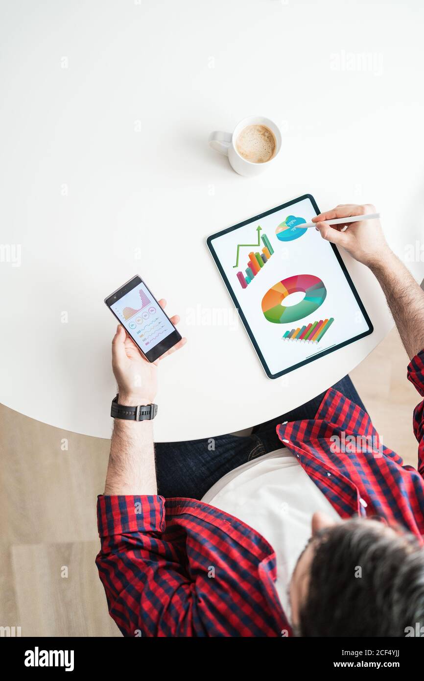 Ein anonymer Mann schreibt auf einem Tablet mit Grafiken. Der Mann arbeitet von zu Hause aus. Der Unternehmer verwendet ein Telefon mit Grafiken von oben Stockfoto