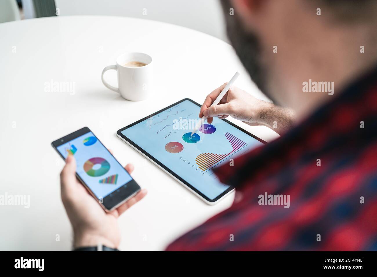 Ein anonymer Mann schreibt auf einem Tablet mit Grafiken. Der Mann arbeitet von zu Hause aus. Der Unternehmer hält ein Telefon mit Grafiken von oben Stockfoto