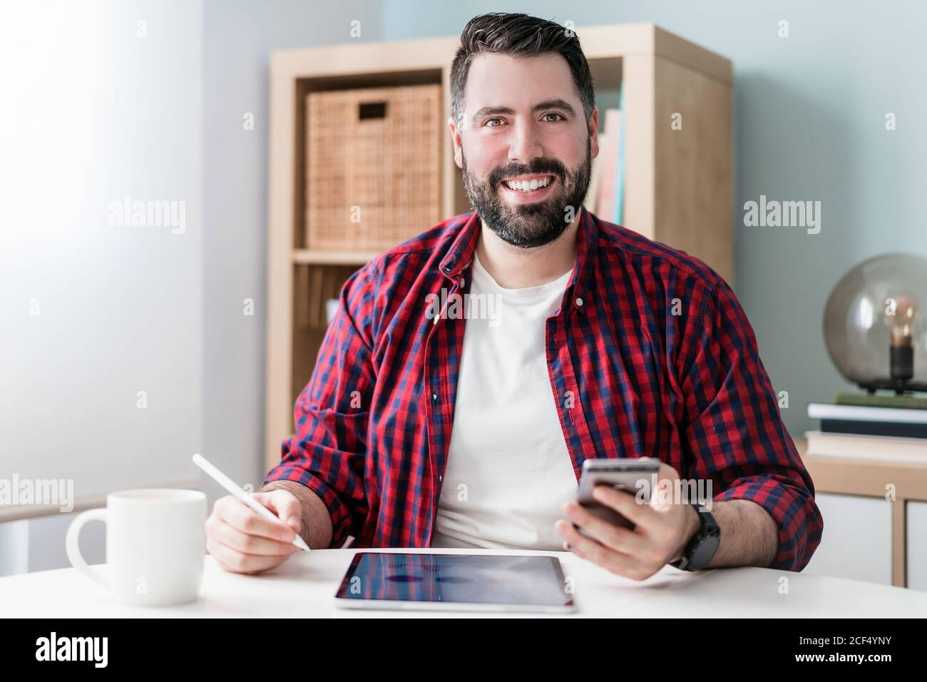 Ein junger Mann schreibt auf einem Tablet. Der Mann arbeitet von zu Hause aus. Der Unternehmer hält ein Telefon Stockfoto