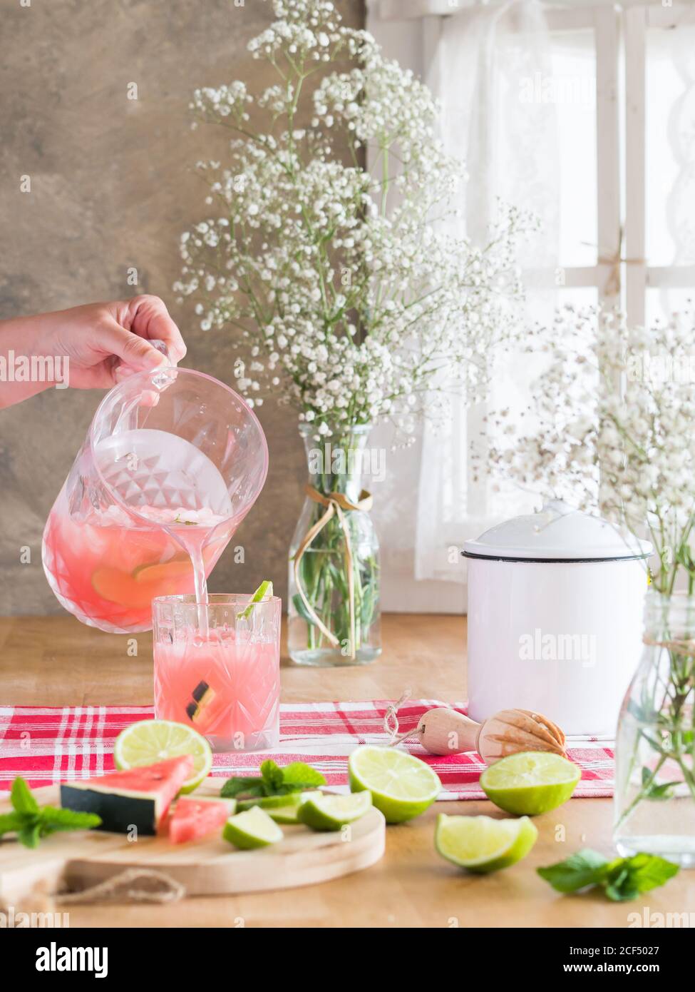 Eisrosa Getränk mit Wassermelone und Limettenscheiben in Gießen Glas aus kaltem Glas auf rustikalem Küchentisch nebenan Bündel von Gypsophila Blumen bei Tageslicht Stockfoto