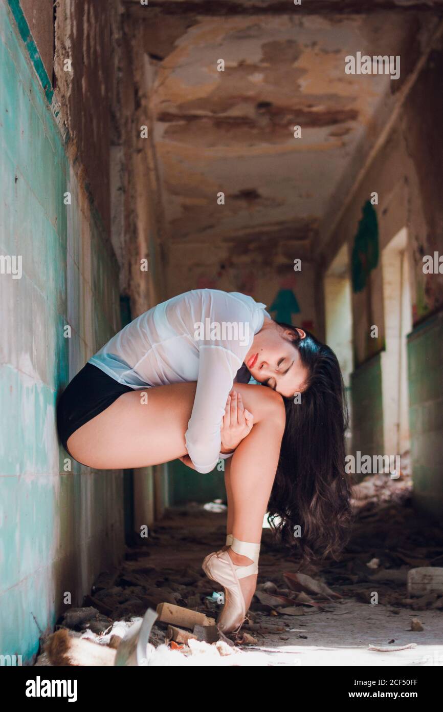 Seitenansicht der schlanken Dame mit geschlossenen Augen umarmen Knie Beim Tanzen im schäbigen Flur eines verlassenen Gebäudes Stockfoto