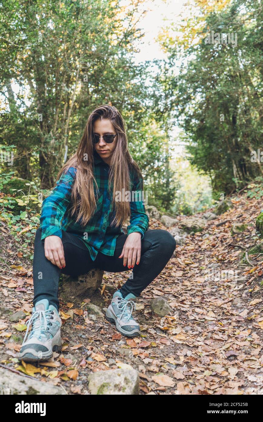 Langhaarige weibliche Hipster in stilvollen lässigen karierten Shirt Sneakers Und trendige Sonnenbrille sitzt auf kleinen Stub auf verspankelt Laubweg zwischen grünen Bäumen Stockfoto