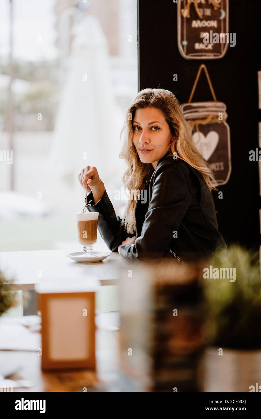 Seitenansicht der langhaarigen trendigen schönen blonden Frau, die aus einem Glas köstlichen schäumenden Kaffee trinkt, während sie die Kamera anschaut Stockfoto