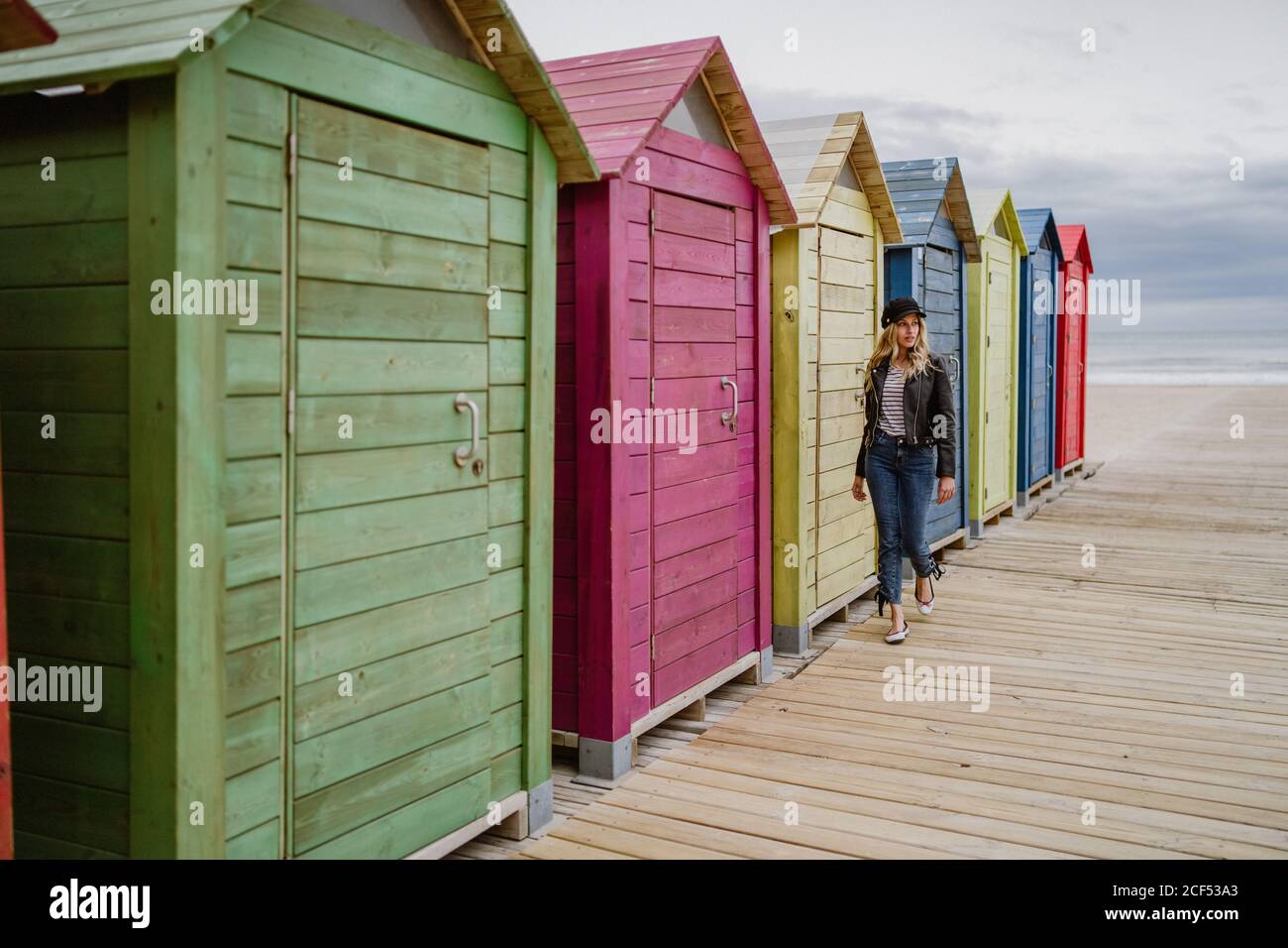 Trendige langhaarige blonde Frau in schwarzer Mütze und Lederjacke lächelt hell an der Kamera und an der hölzernen Strandhütte vorbei Stockfoto