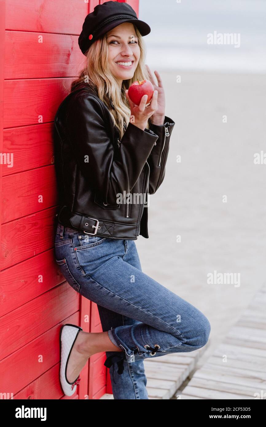 Trendige langhaarige blonde Frau in schwarzer Mütze und Lederjacke lächelt  hell auf die Kamera, die einen roten Apfel isst, während sie sich auf eine  hölzerne Strandhütte stützt Stockfotografie - Alamy