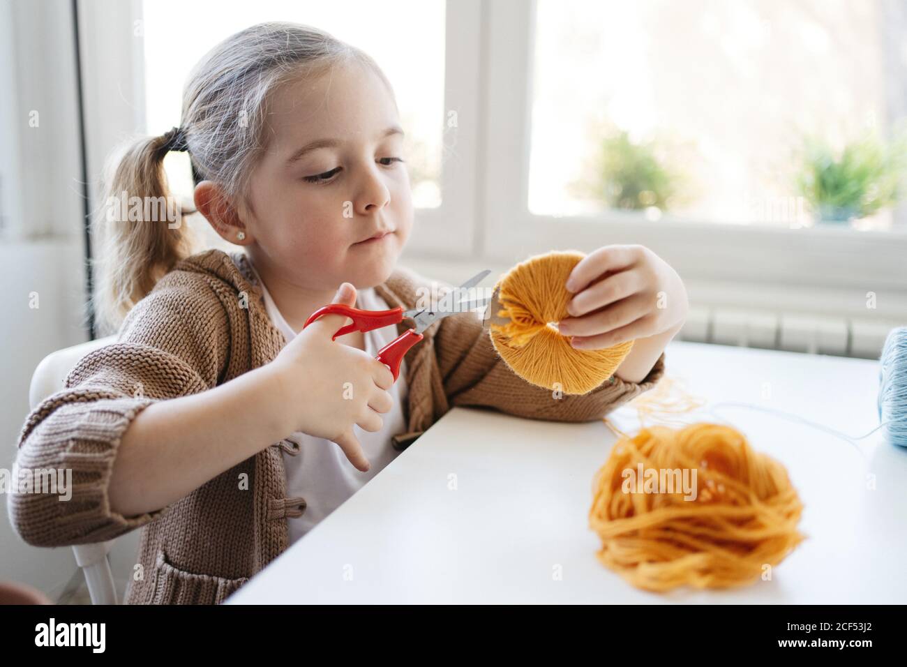 Entzückendes Kind in legerer Kleidung mit orangefarbenem Muster aus Wolle Fäden und Schneiden mit einer Schere zwischen den Kreisen während der Arbeit Kreatives Handwerk in der Werkstatt Stockfoto
