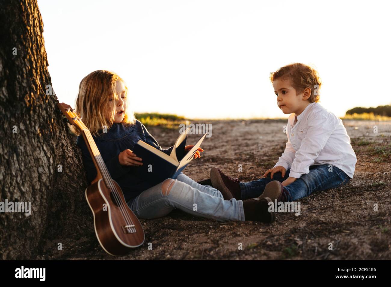 Seitenansicht von liebenswert kleines Mädchen lesen interessante Geschichte zu Jüngerer Bruder, während er zusammen unter dem Baum mit Ukulele-Gitarre sitzt Im Sommer Tag auf dem Land Stockfoto