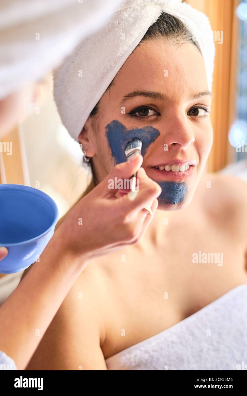 Anonyme Frau, die während des Eingriffs zu Hause eine blaue Tonmaske auf das Gesicht einer fröhlichen Frau anwendet, die die Kamera in einem weißen Handtuch anschaut Stockfoto