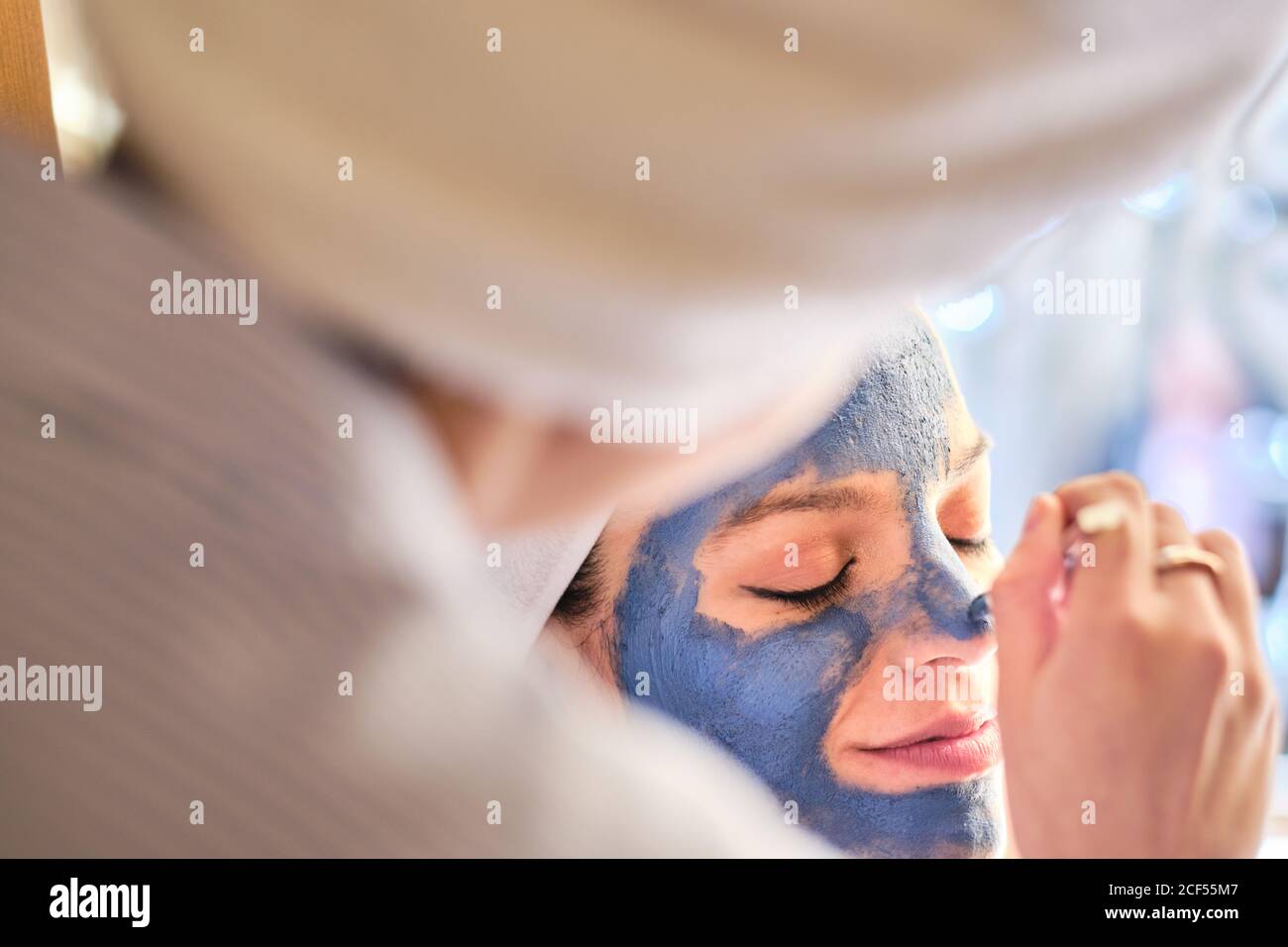 Rückansicht einer anonymen Frau, die während des Eingriffs zu Hause eine blaue Tonmaske auf das Gesicht einer ruhigen Frau mit geschlossenen Augen in einem weißen Handtuch anwendet Stockfoto