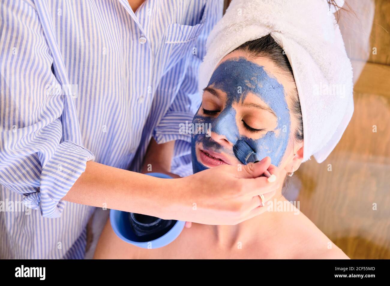 Seitenansicht einer anonymen Frau, die während des Eingriffs zu Hause eine blaue Tonmaske auf das Gesicht einer ruhigen Frau mit geschlossenen Augen in einem weißen Handtuch anwendet Stockfoto