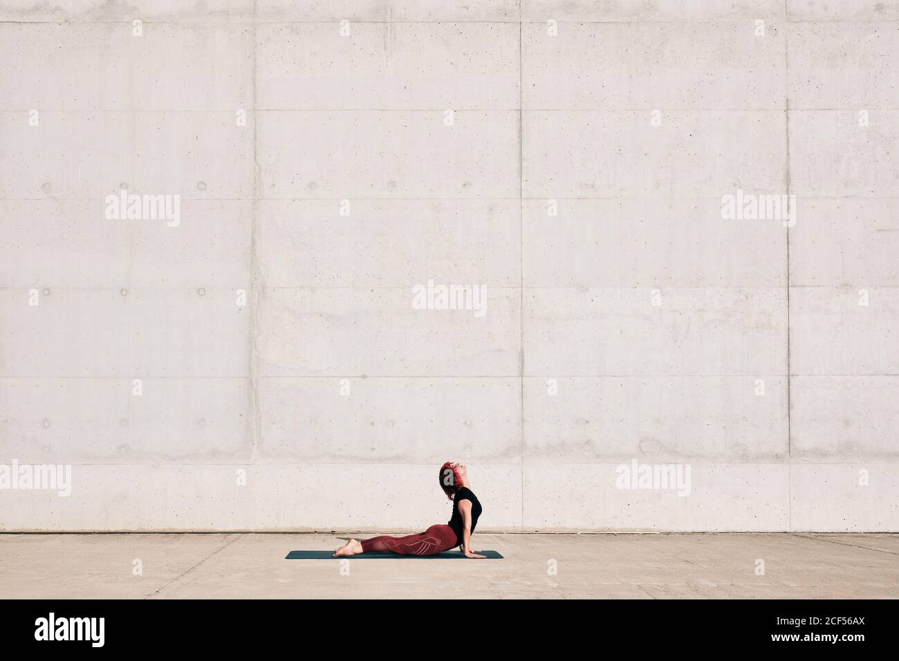 Seitenansicht der trendigen Sportlerin im Sportswear-Bereich Baby Cobra Yoga-Übung auf Sportmatte während des Trainings allein An sonnigen Tagen auf der Straße gegen Betonmauer Stockfoto