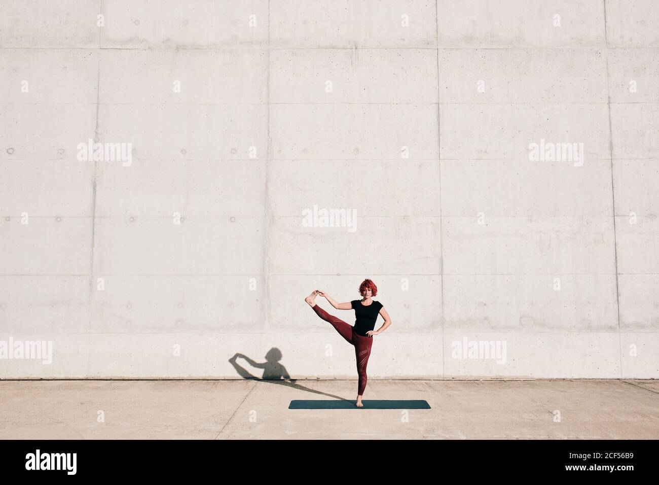 Trendy fit weibliche Sportlerin in Sportswear tun ausgestreckte Hand zu Big Toe Yoga Pose auf Sportmatte beim Training allein An sonnigen Tagen auf der Straße gegen Betonmauer Stockfoto