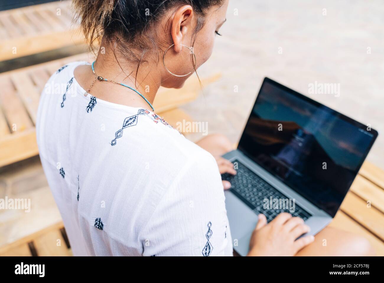 Rückansicht Frau in legerer Kleidung, die ihren Laptop durchstöbert, während sie mit gekreuzten Beinen auf einem hölzernen Liegestuhl auf der tropischen Terrasse sitzt Stockfoto