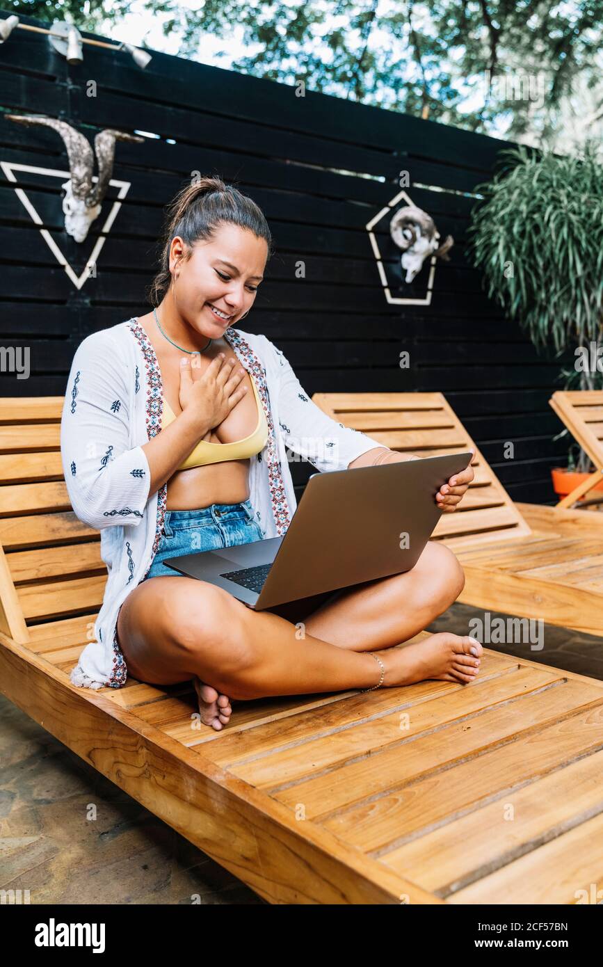Fröhliche Frau in legerer Kleidung, die auf einem Laptop mit gekreuzten Beinen auf einem Holzdeckelstuhl auf der tropischen Terrasse sitzt Stockfoto