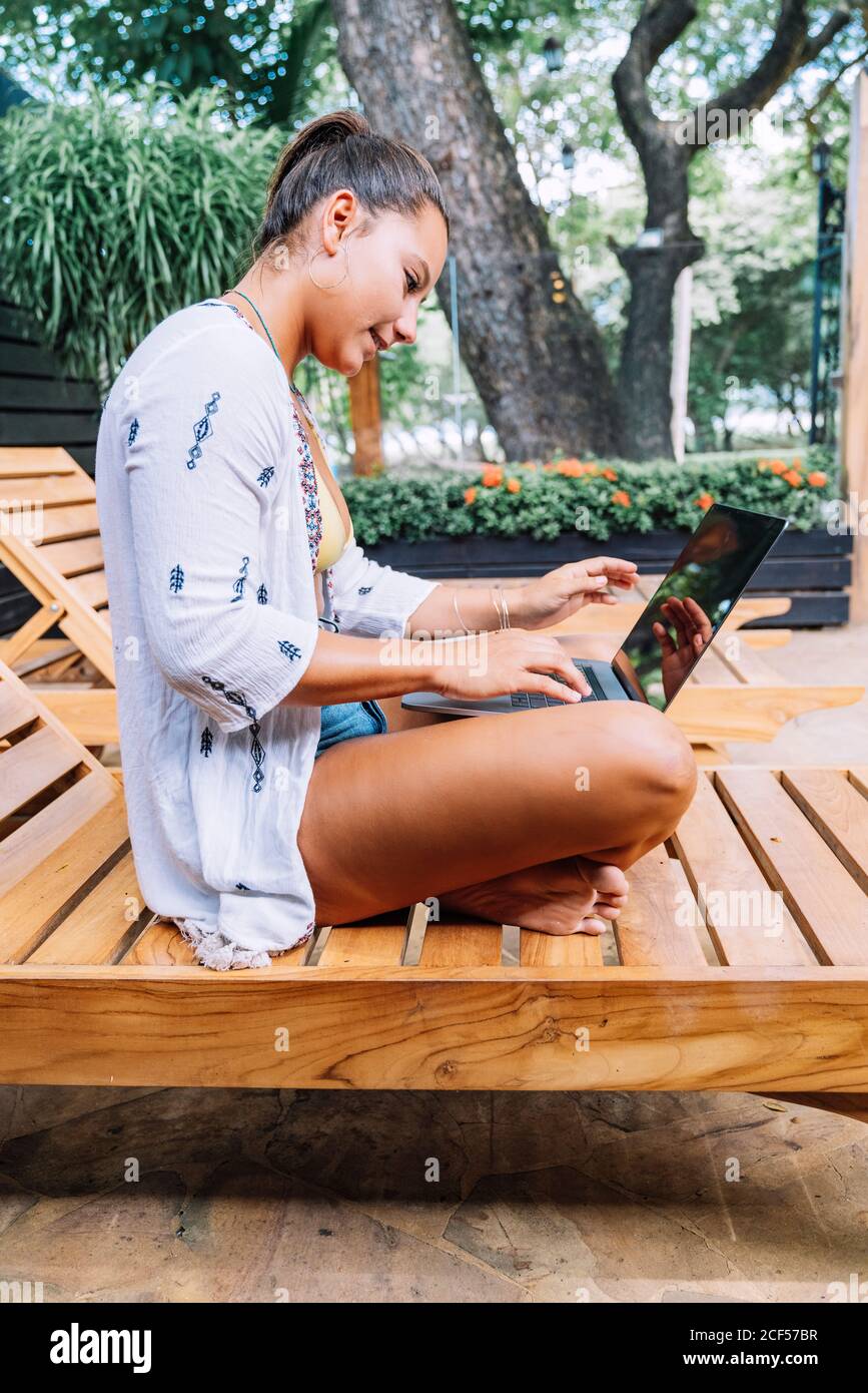 Seitenansicht einer Frau in legerer Kleidung, die ihren Laptop durchstöbert, während sie mit gekreuzten Beinen auf einem Holzdeckelstuhl auf der tropischen Terrasse sitzt Stockfoto