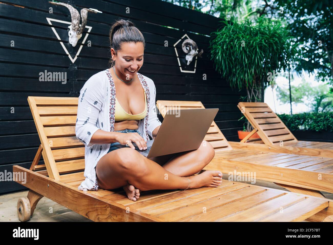 Fröhliche Frau in legerer Kleidung, die auf einem Laptop mit gekreuzten Beinen auf einem Holzdeckelstuhl auf der tropischen Terrasse sitzt Stockfoto