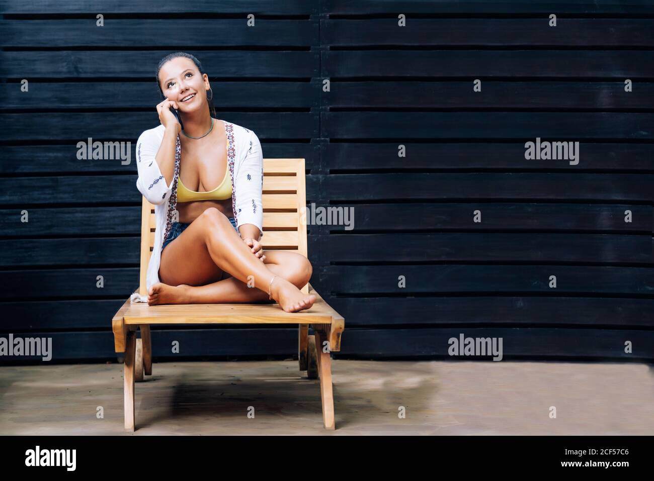 Glückliche Frau in legerer Kleidung, die auf dem Smartphone spricht, während sie mit gekreuzten Beinen auf einem Holzdeckelstuhl auf der tropischen Terrasse in Costa Rica sitzt Stockfoto