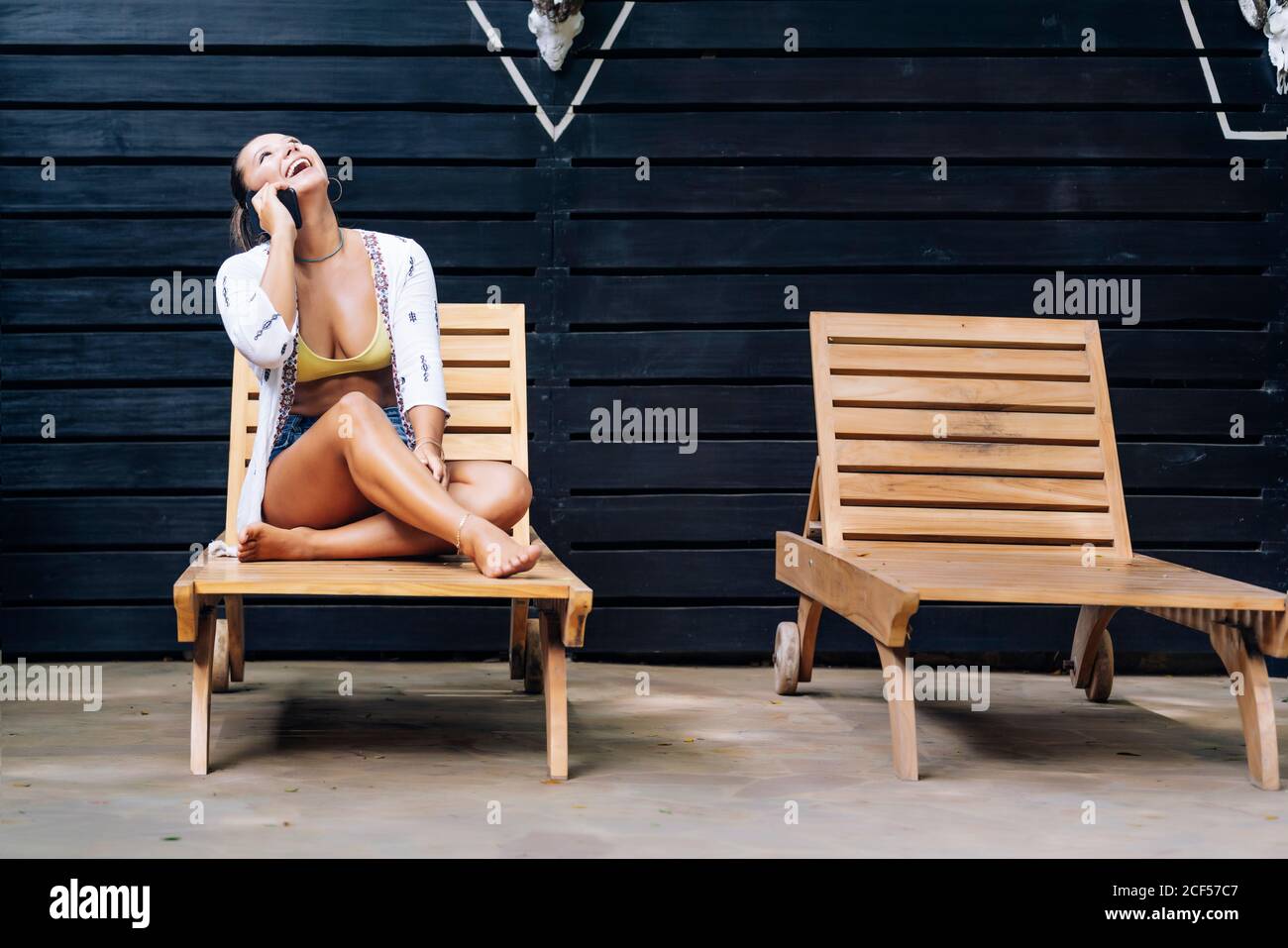 Glückliche Frau in legerer Kleidung, die auf dem Smartphone spricht, während sie mit gekreuzten Beinen auf einem Holzdeckelstuhl auf der tropischen Terrasse in Costa Rica sitzt Stockfoto