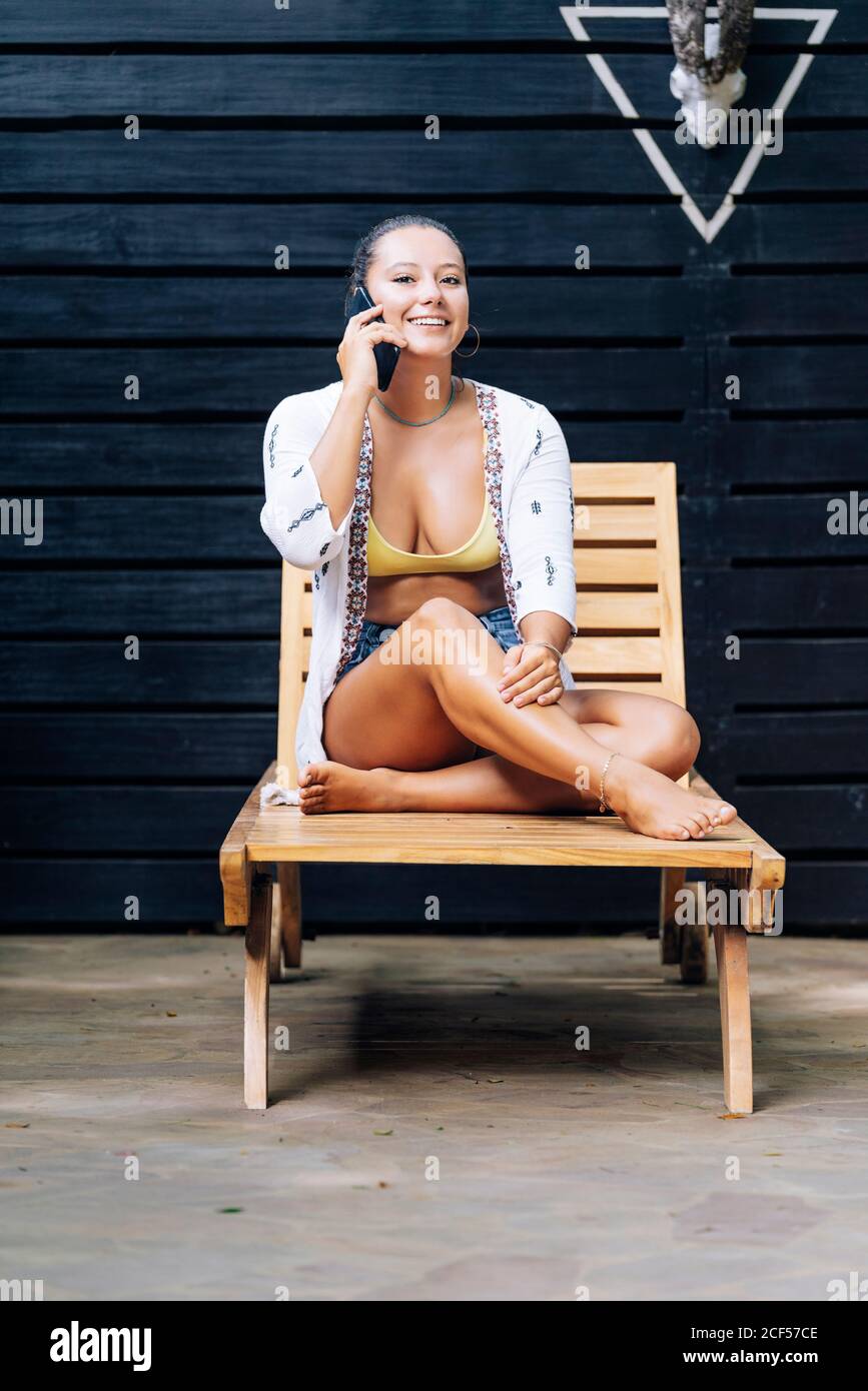 Glückliche Frau in legerer Kleidung, die auf dem Smartphone mit gekreuzten Beinen auf einem Holzdeckelstuhl auf der tropischen Terrasse in Costa Rica Lippe beißt und spricht Stockfoto