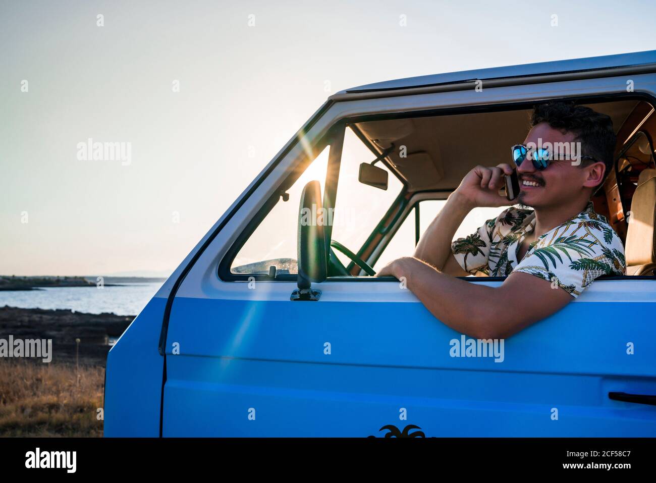 Seitenansicht des lächelnden erwachsenen Mannes in Sonnenbrillen und lässig Kleidung, die wegschaut und telefonieren kann, während man im Sitzen sitzt Blauer Van an sonniger Abendküste Stockfoto