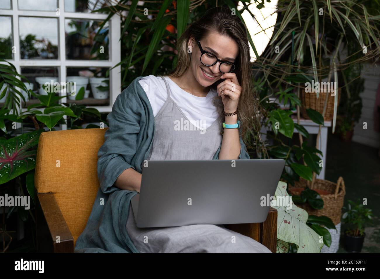 Nahaufnahme Porträt der lächelnden Gärtnerin in Gläsern Tragen Sie Leinenkleid, sitzen auf einem Stuhl im grünen Haus, mit Laptop und sprechen im Web-Anruf Stockfoto