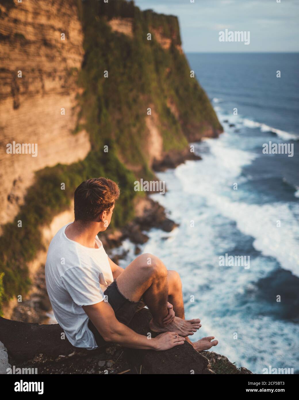 Seitenansicht des Mannes, der allein auf einer Klippe in der Höhe sitzt und die majestätische Landschaft der Meereswellen, Bali, betrachtet Stockfoto