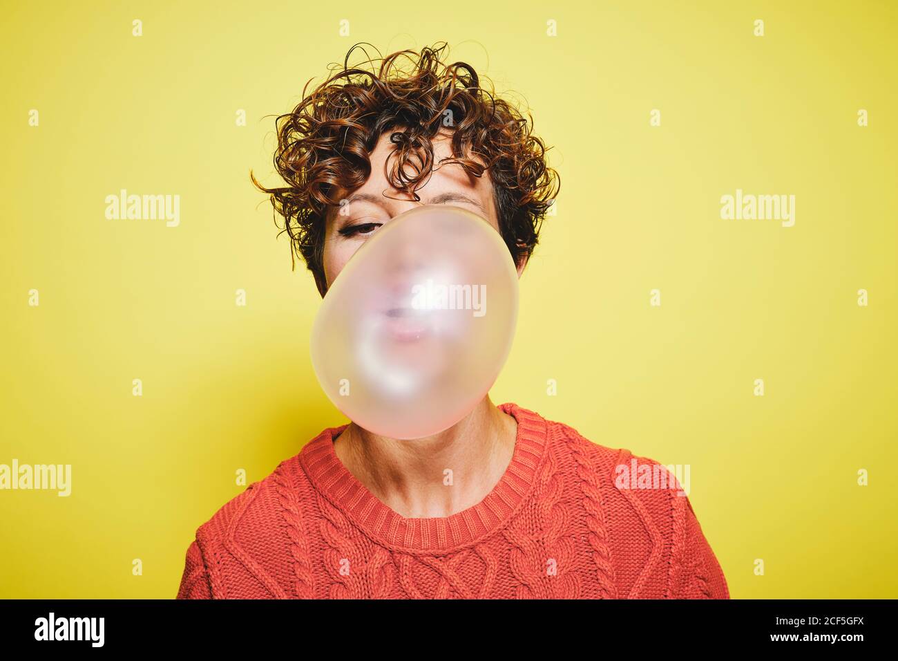 Junge lockig behaarte Frau in orange Pullover weht Blase Gummi Während Sie vor gelbem Hintergrund stehen Stockfoto