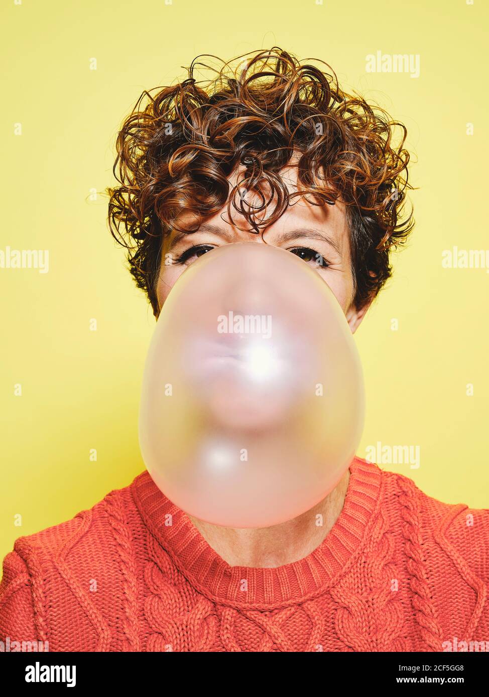 Junge lockig behaarte Frau in orange Pullover weht Blase Gummi Blick auf die Kamera, während Sie vor gelbem Hintergrund stehen Stockfoto
