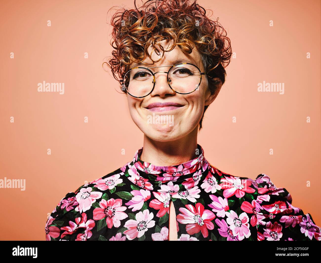 Glücklich junge lockig behaart weiblich in stilvollen floral Bluse und Trendige Brillen lächeln und vor rotem Hintergrund auf die Kamera schauen Stockfoto