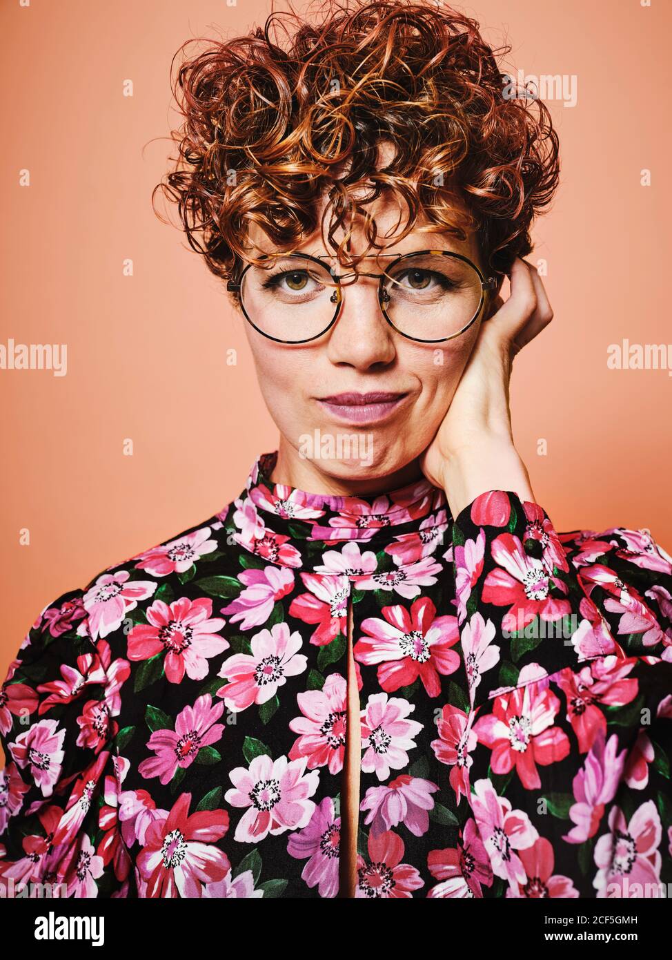 Nachdenklich zweifelhaft schöne lockige behaarte Weibchen in trendigen Brillen und Stilvolle bunte Bluse mit floralem Ornament Blick auf Kamera gegen Rosa Hintergrund Stockfoto