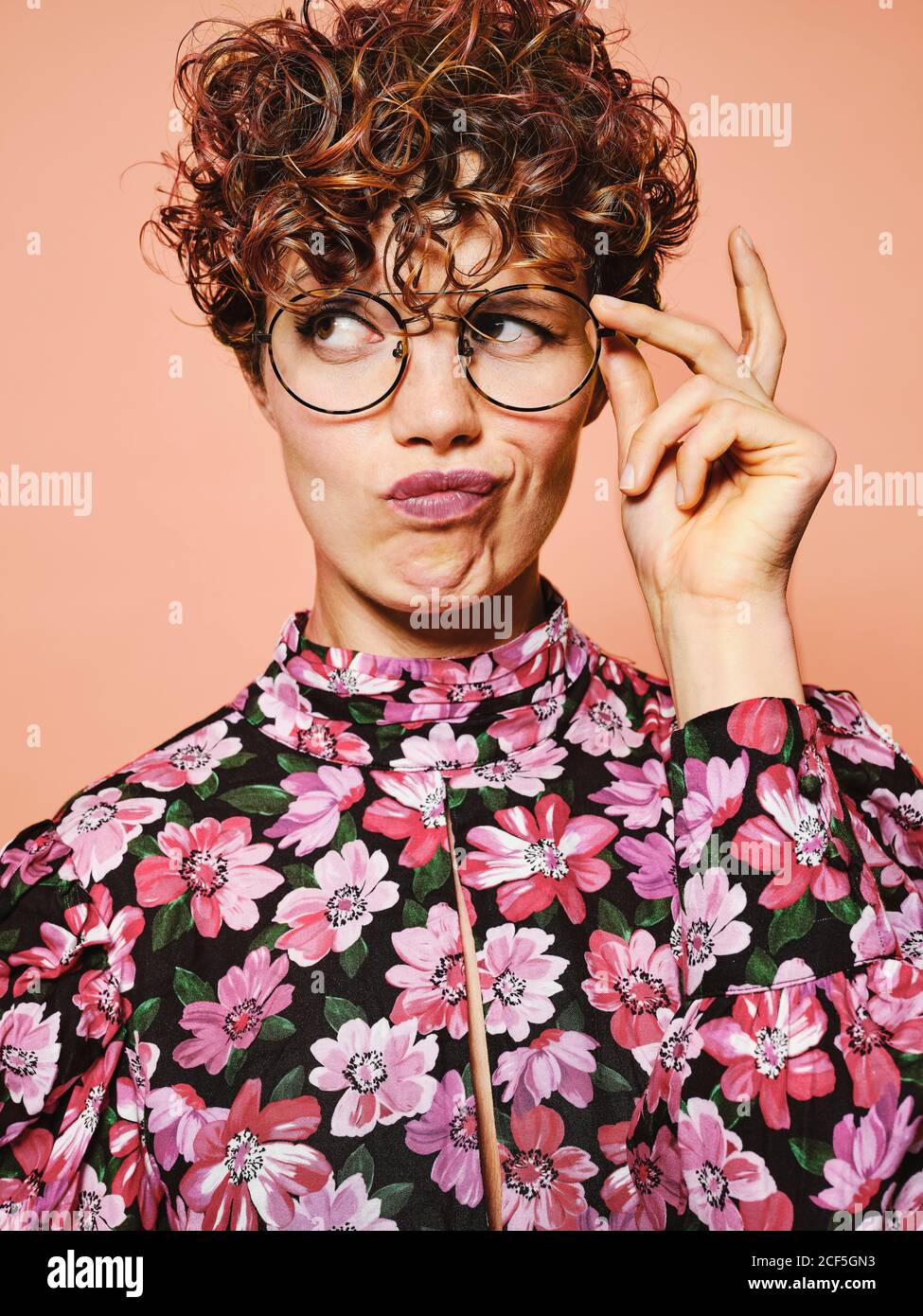 Nachdenklich zweifelhaft schöne lockige behaarte Weibchen in trendigen Brillen und Stylische, farbenfrohe Bluse mit floralem Ornament, das gegen Rosa abblickt Hintergrund Stockfoto