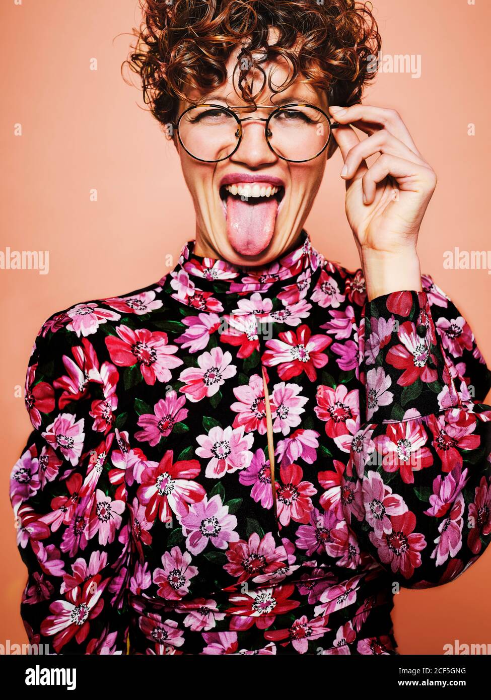 Schöne Lockenhaarige Weibchen in trendigen Brillen und stilvolle bunte Bluse mit floralen Ornament Blick auf Kamera und macht lustig Grimace mit Zunge aus stehen vor rosa Hintergrund Stockfoto