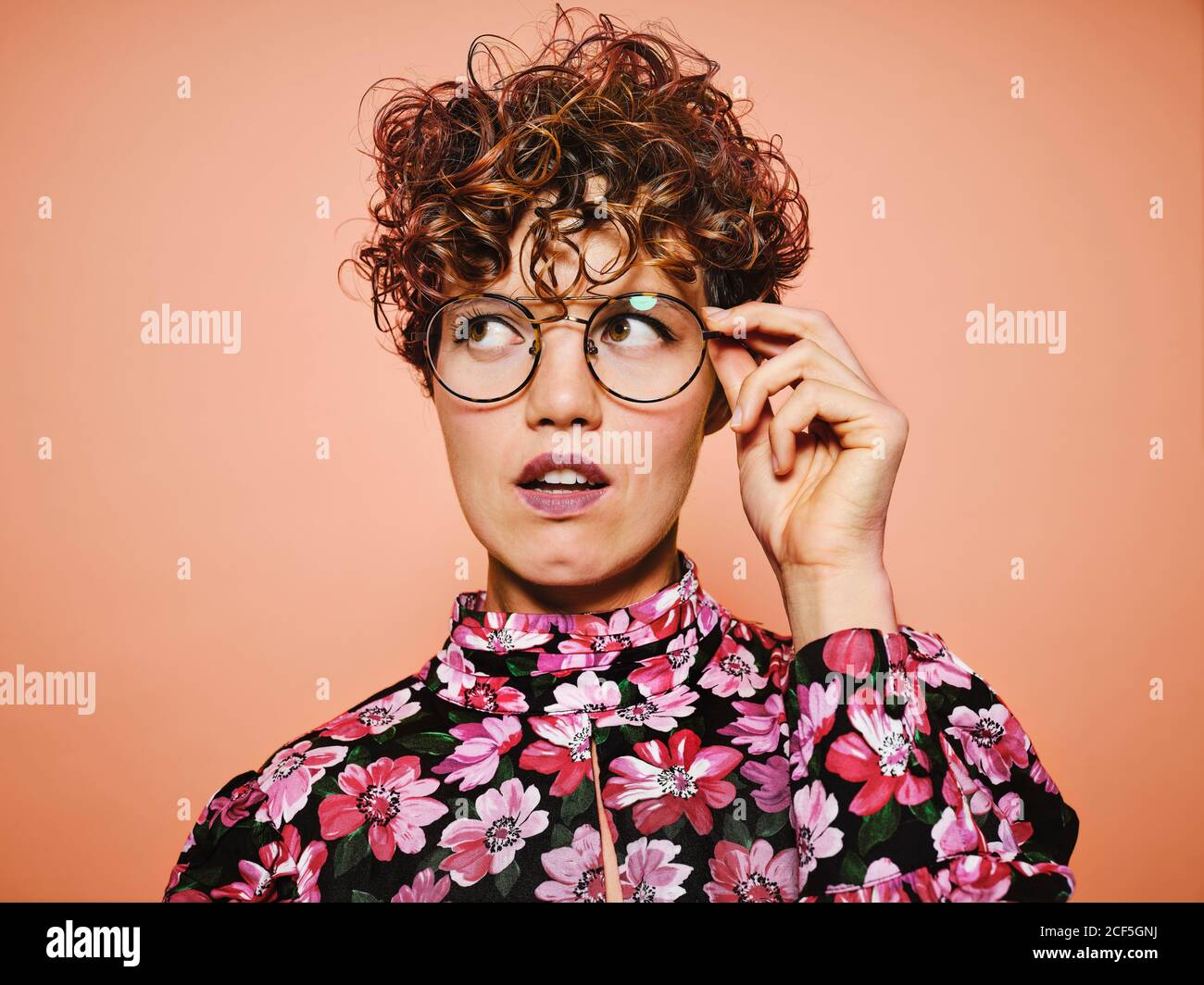 Nachdenklich zweifelhaft schöne lockige behaarte Weibchen in trendigen Brillen und Stylische, farbenfrohe Bluse mit floralem Ornament, das gegen Rosa abblickt Hintergrund Stockfoto