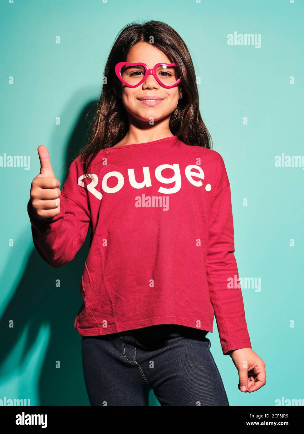 Glücklich präteen Mädchen in rosa Herzform Gläser und lässig Kleidung lächelt an der Kamera mit Daumen nach oben stehen gegen türkis Hintergrund im modernen Studio Stockfoto