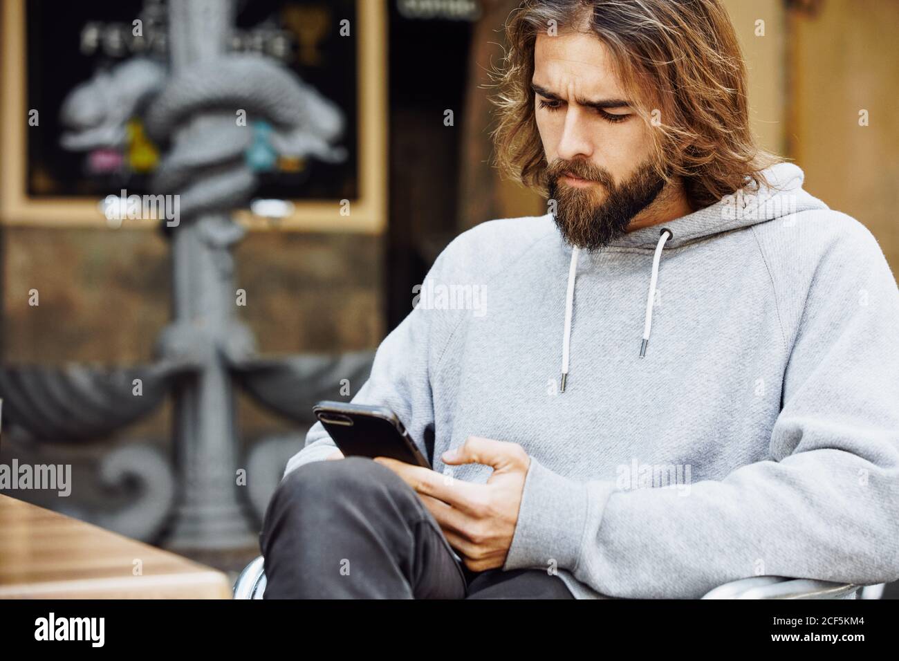 Junger bärtiger hübscher Mann im lässigen Pullover, der im Café sitzt Konzentriert sich auf das Mobiltelefon Stockfoto
