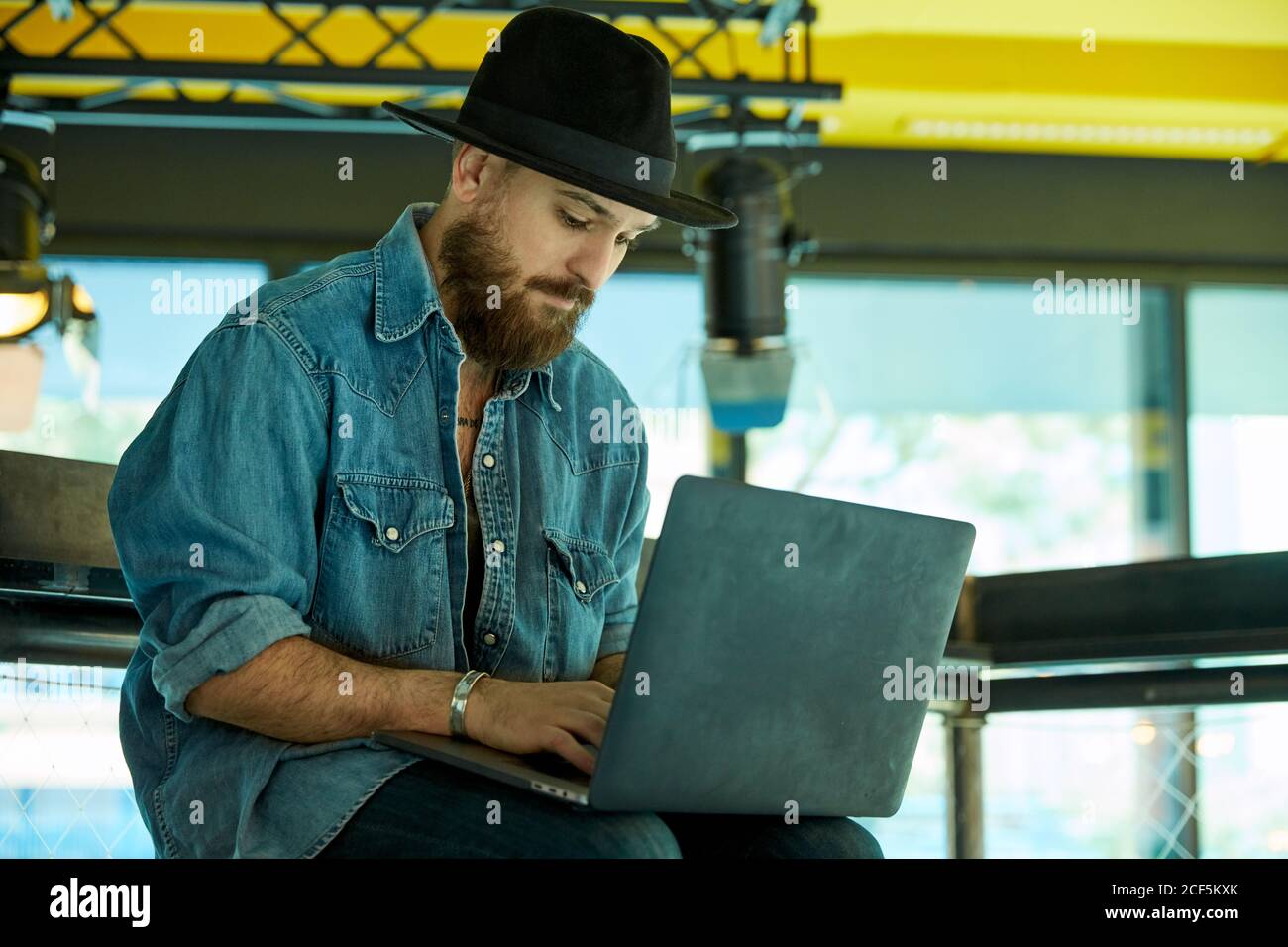 Junger bärtiger hübscher Mann mit schwarzem Hut sitzend und arbeitend Notebook auf Runden halten Stockfoto