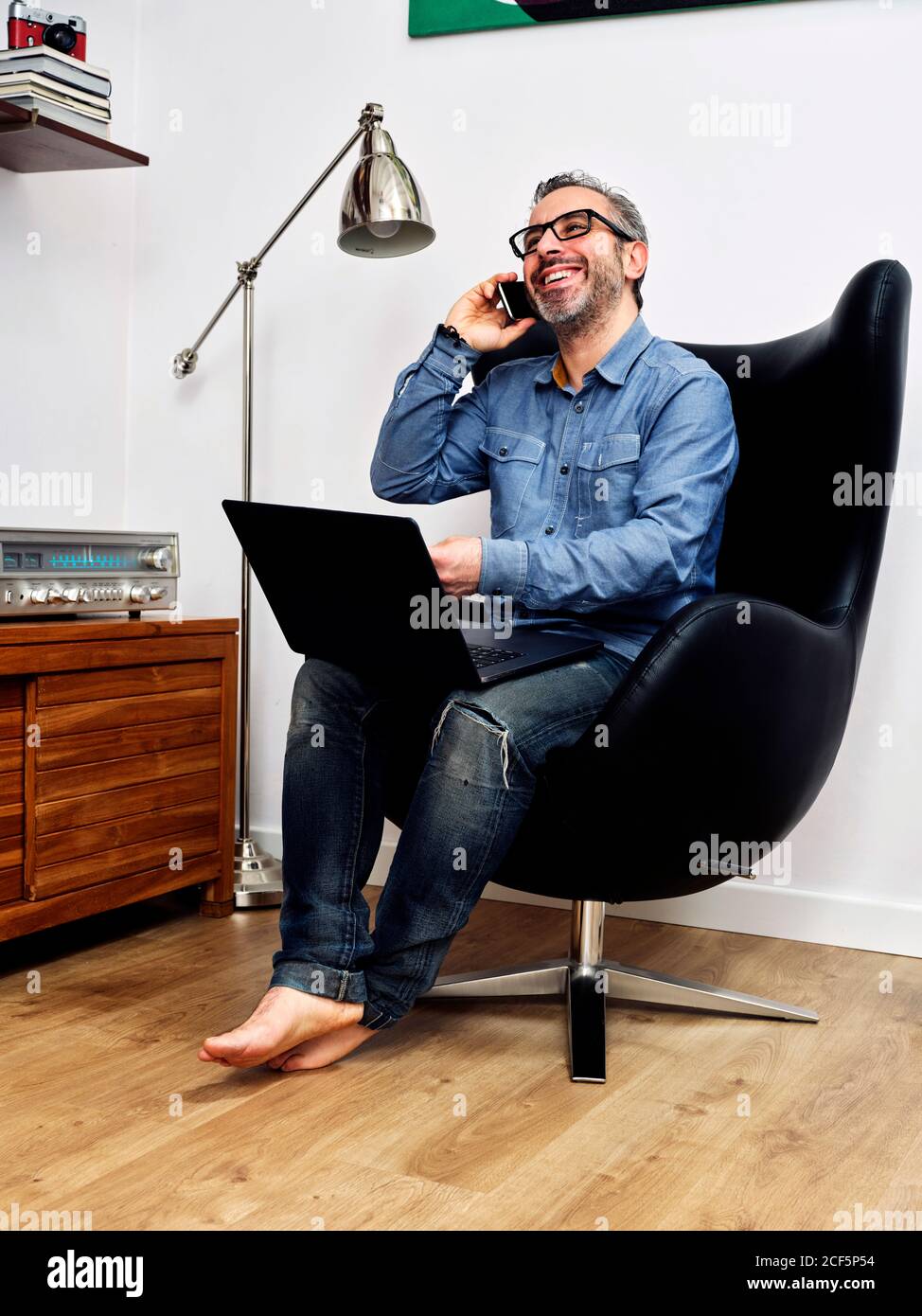 Mann, der am Laptop arbeitet und auf dem Handy spricht Telefon lächelnd mit einem entspannten Kaffee im Sessel an Zu Hause Stockfoto