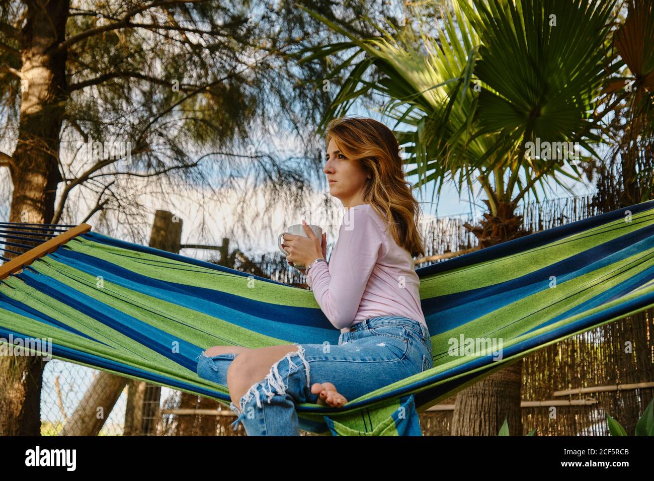 Ruhige Frau im lässigen Outfit sitzt in der Hängematte mit Tasse Von heißen Getränken im Innenhof und genießen Sommerurlaub Stockfoto
