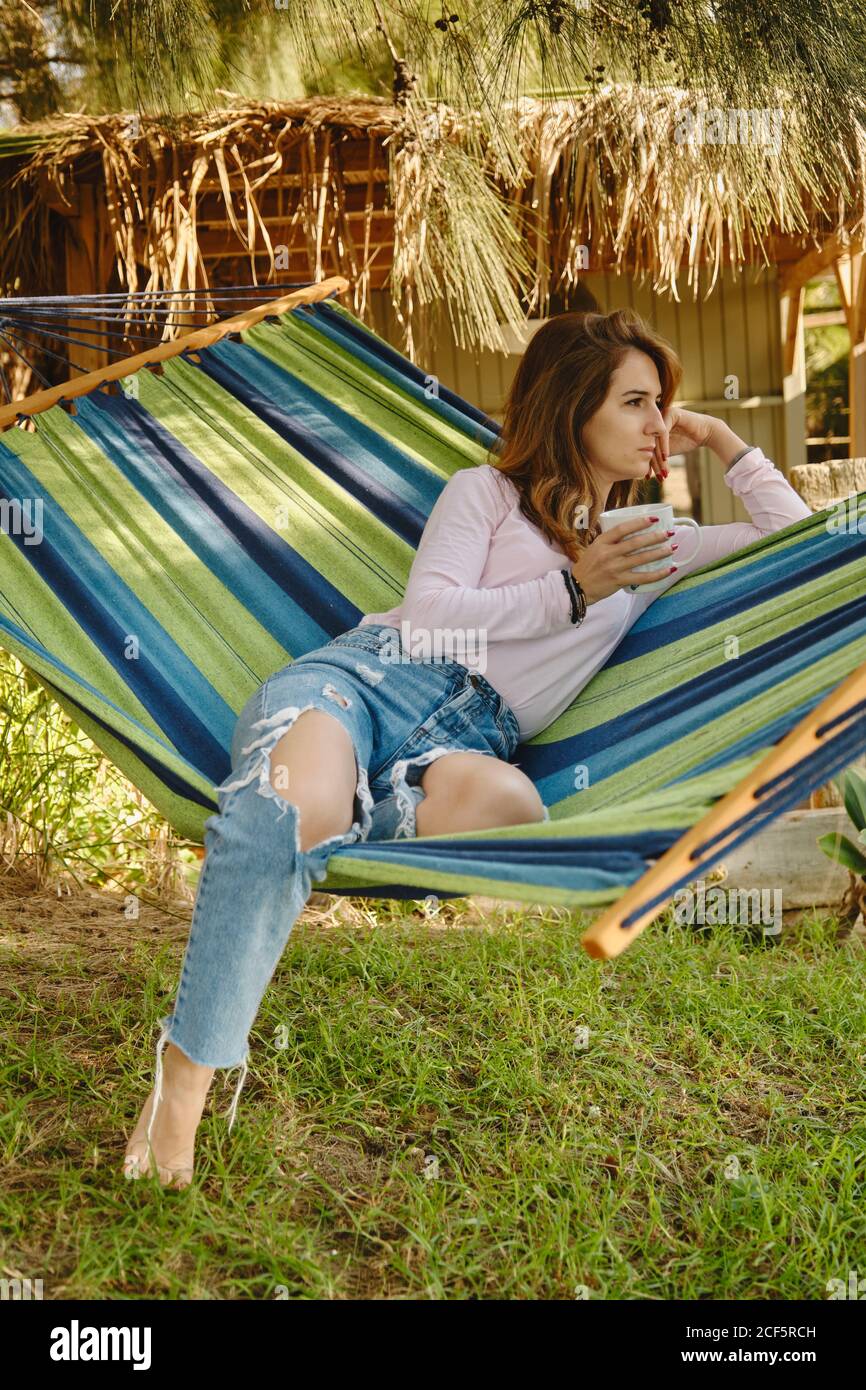 Ruhige Frau im lässigen Outfit sitzt in der Hängematte mit Tasse Von heißen Getränken im Innenhof und genießen Sommerurlaub Stockfoto