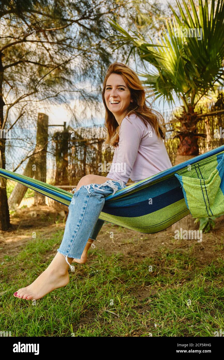 Lächelnd ruhige Frau im lässigen Outfit, das in der Hängematte innen sitzt Innenhof und genießen Sommerurlaub Stockfoto