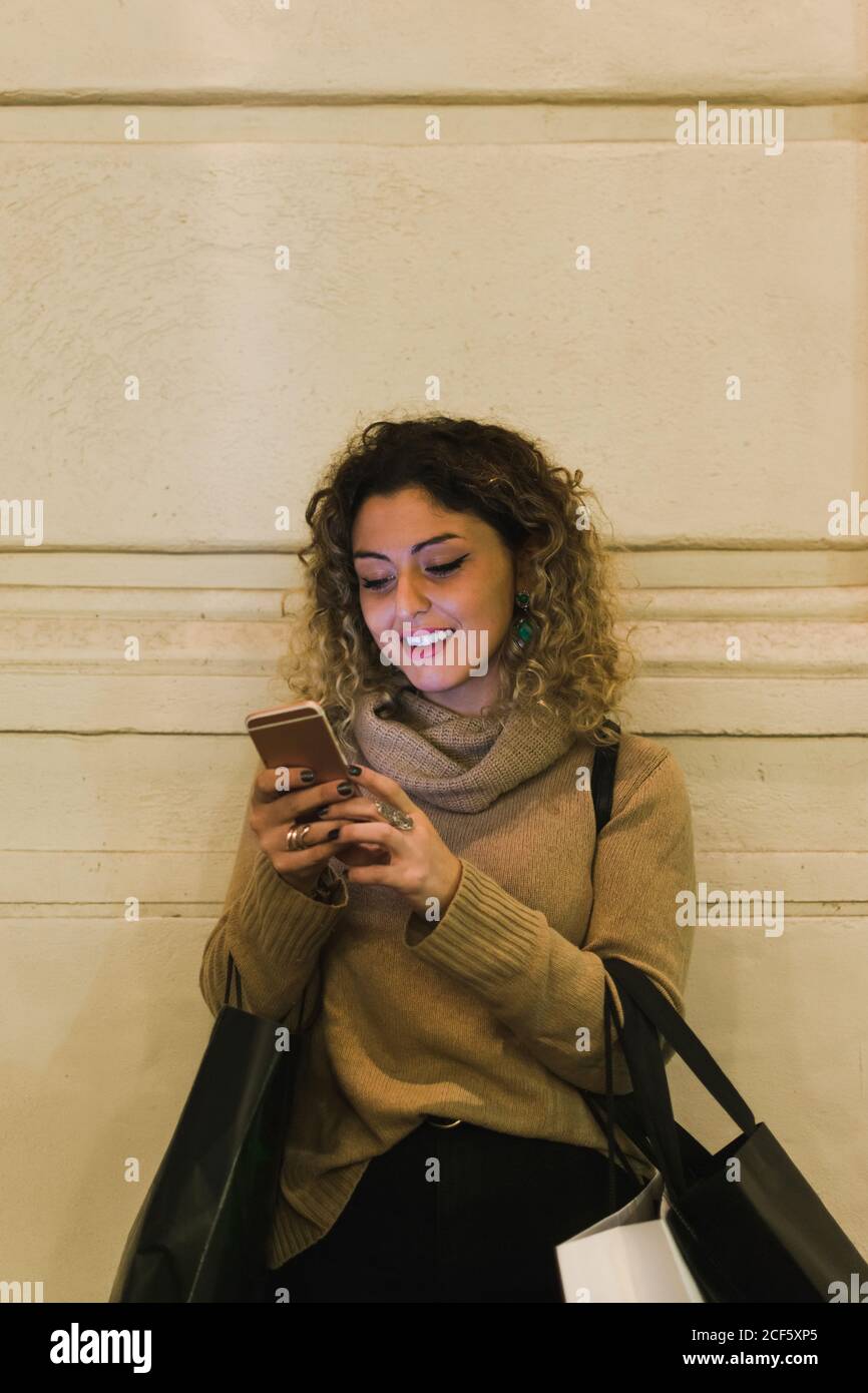Zufriedene, lockige junge Frau in legerer Kleidung, die lächelt, während sie das Mobiltelefon mit Einkaufstaschen an der Wand anlehnt Stockfoto