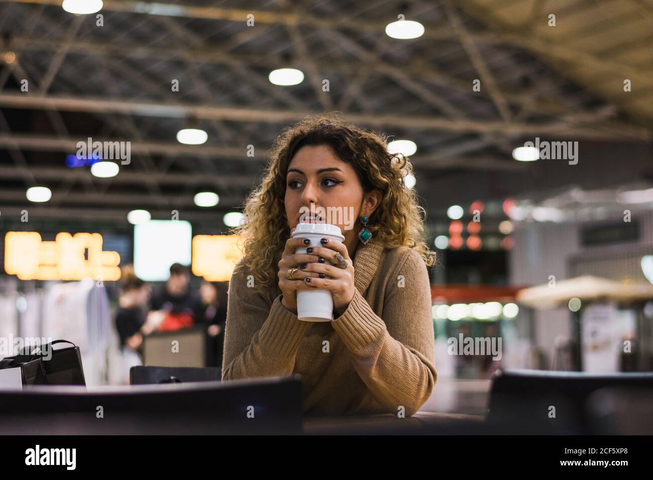 Zeitgemäße Dame in beige Pullover lehnte Ellbogen auf Tisch Mit Kaffee in Flughafen auf verschwommenem Hintergrund zu gehen Stockfoto