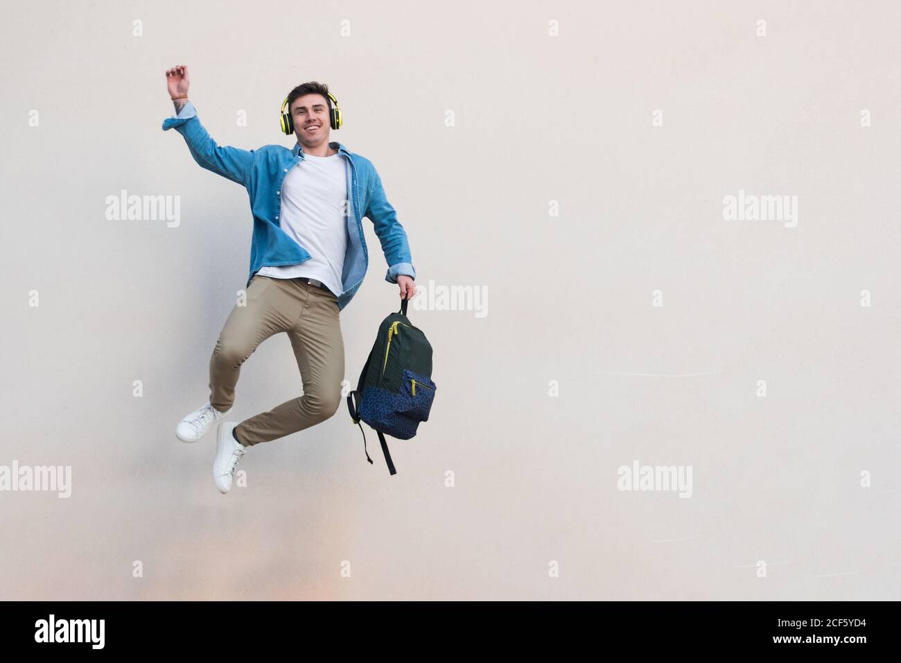Fröhliche stilvolle Mann Kopfhörer mit Rucksack heben Hand feiern Sieg Und springende umklammernden Fuß auf Hintergrund der grauen Wand in Sonniger Tag Stockfoto