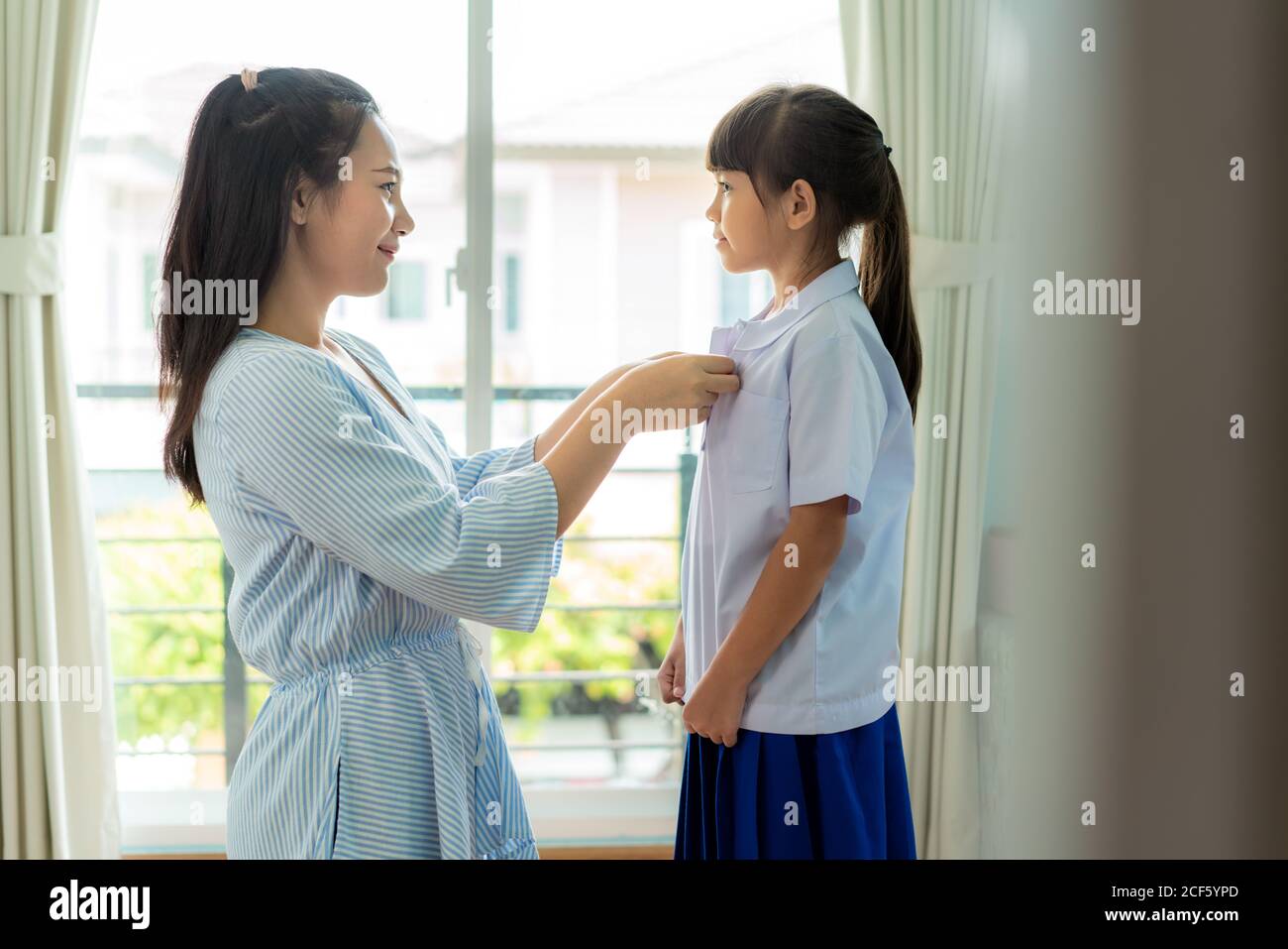 Asiatische Mutter Vorbereitung Grundschüler Uniform zu ihrem kleinen Tochter für die Schule im Wohnzimmer zu Hause. Die morgendliche Schulroutine für den Tag in der LIF Stockfoto