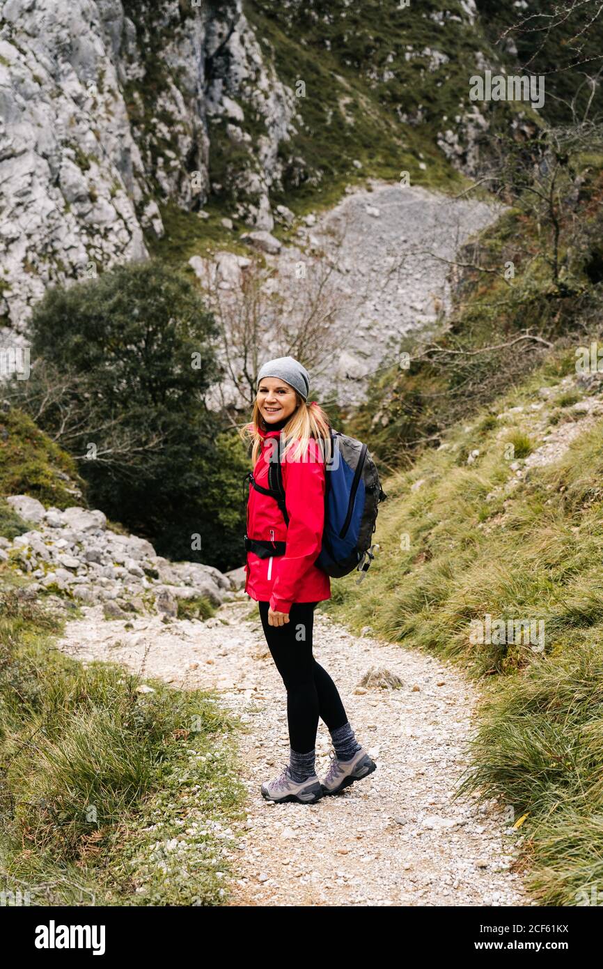 Seitenansicht einer aktiven Wandererin in roter Jacke mit schwerem Rucksack, die die Kamera anschaut, während sie in den Bergen in den Gipfeln Europas, Asturias, Spanien, läuft Stockfoto