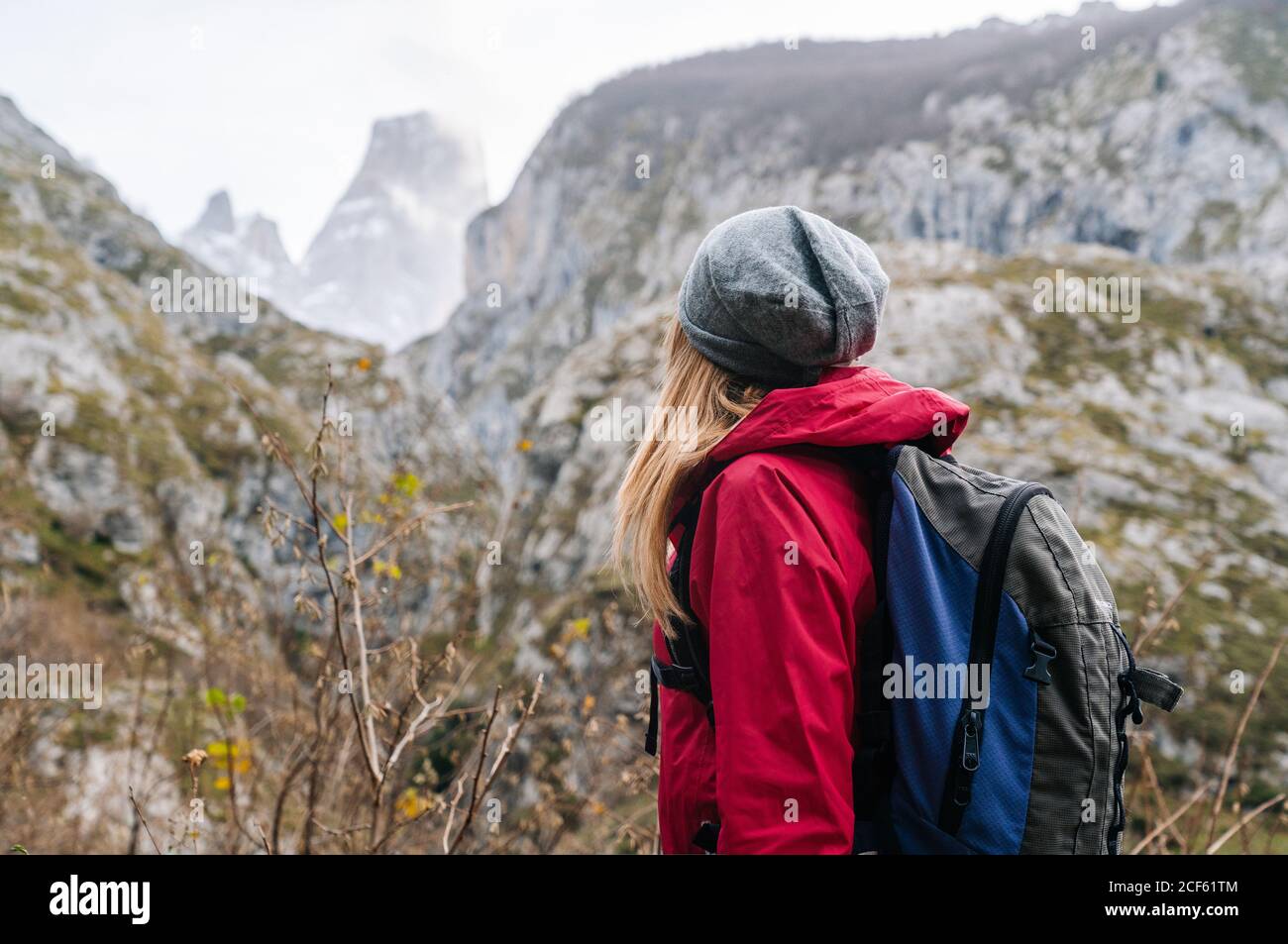Seitenansicht einer nicht erkennbaren aktiven Wandererin in roter Jacke mit schwerem Rucksack, die auf die Berge in den Gipfeln Europas, Asturiens, Spaniens blickt Stockfoto