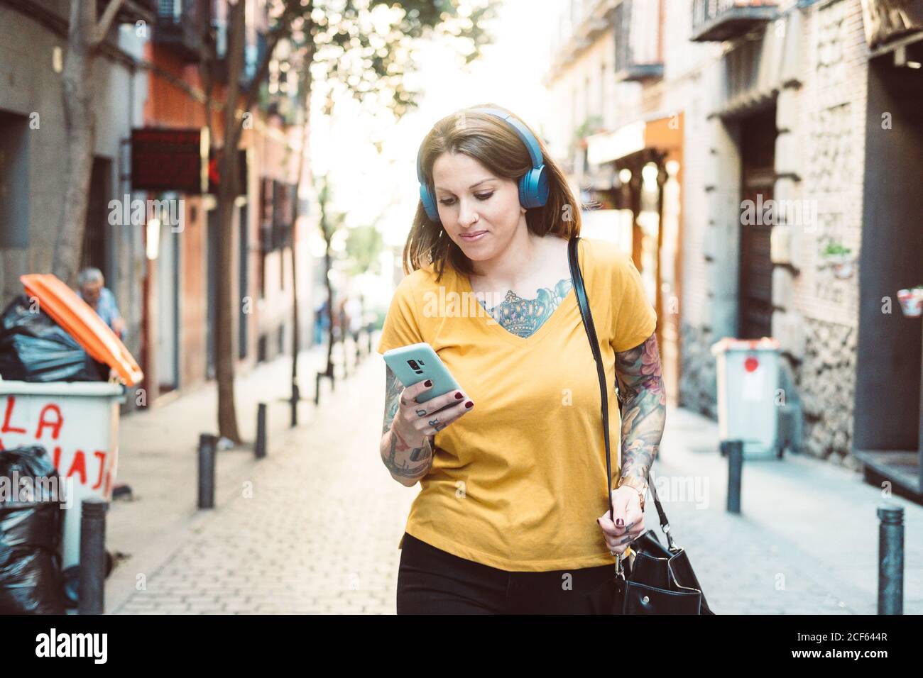 Aufmerksamer Hipster tätowierte eine Frau in Kopfhörern mit dem Smartphone, während sie auf der sonnigen Stadtstraße stand Stockfoto