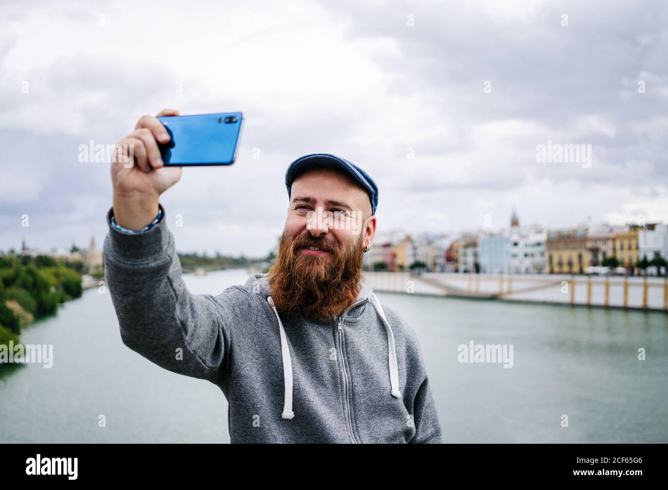 Content männliche Reisende in lässiger Kleidung lächeln und Selfie machen Und Friedenszeichen auf dem Handy, während man auf schaukelte Zaun des Kais Stockfoto