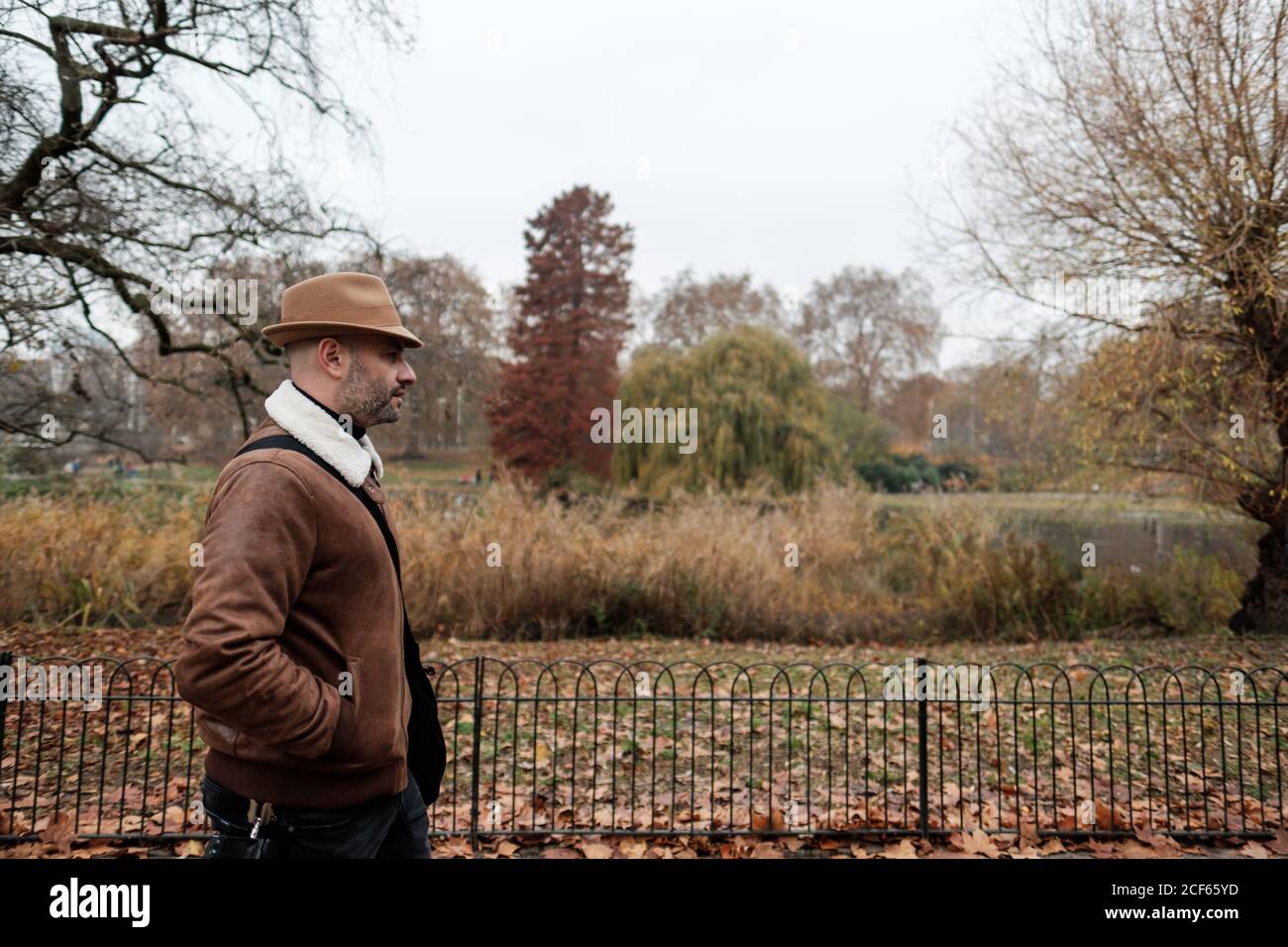 Seitenansicht eines erwachsenen Mannes in legerem Outfit, der die Hände hält In Taschen zu Fuß in der Nähe von Zaun in ruhigen Herbst Park in London Stockfoto